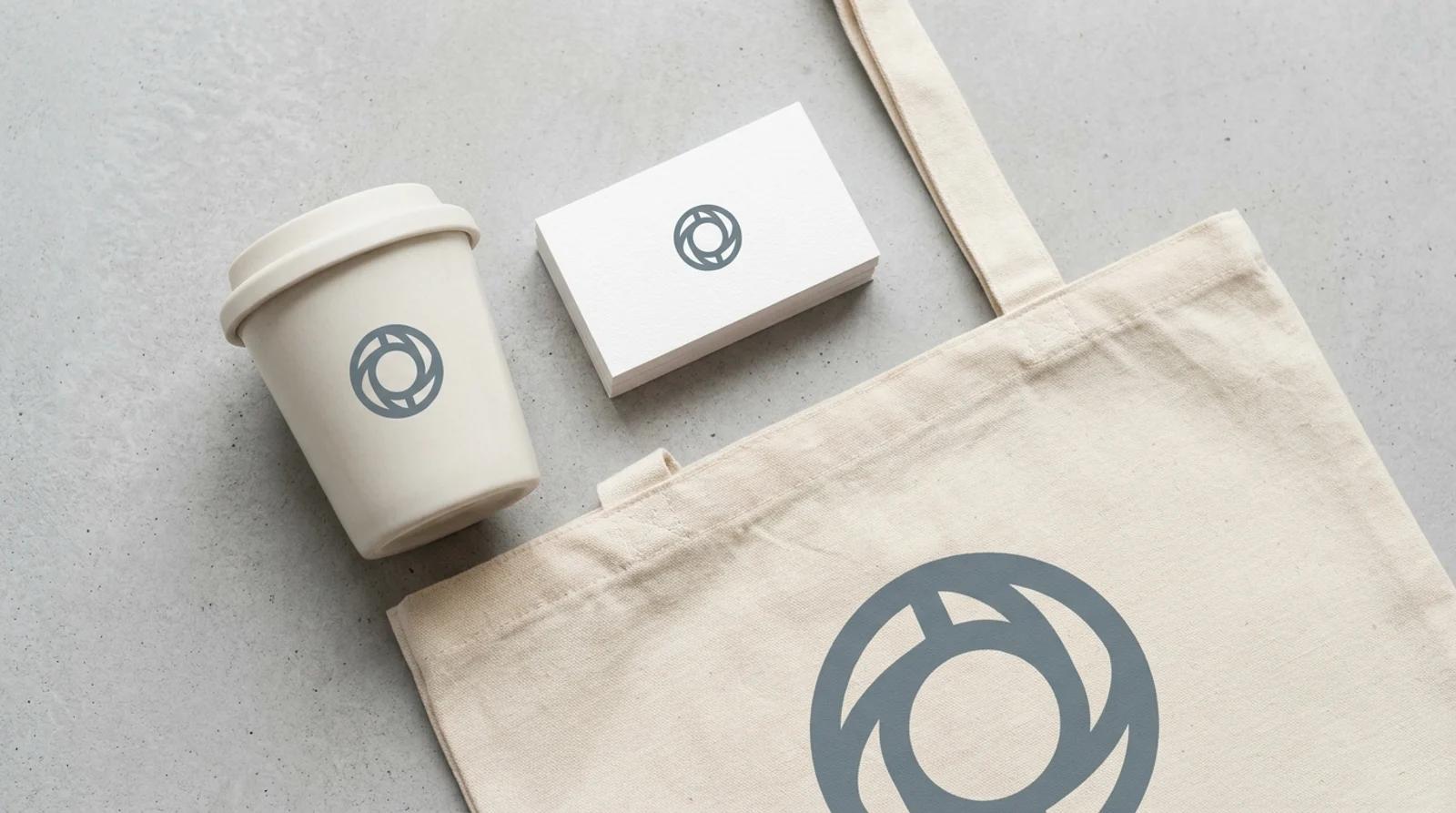 A brand mockup showing a simple icon mark on a business card, a coffee cup, and a tote bag